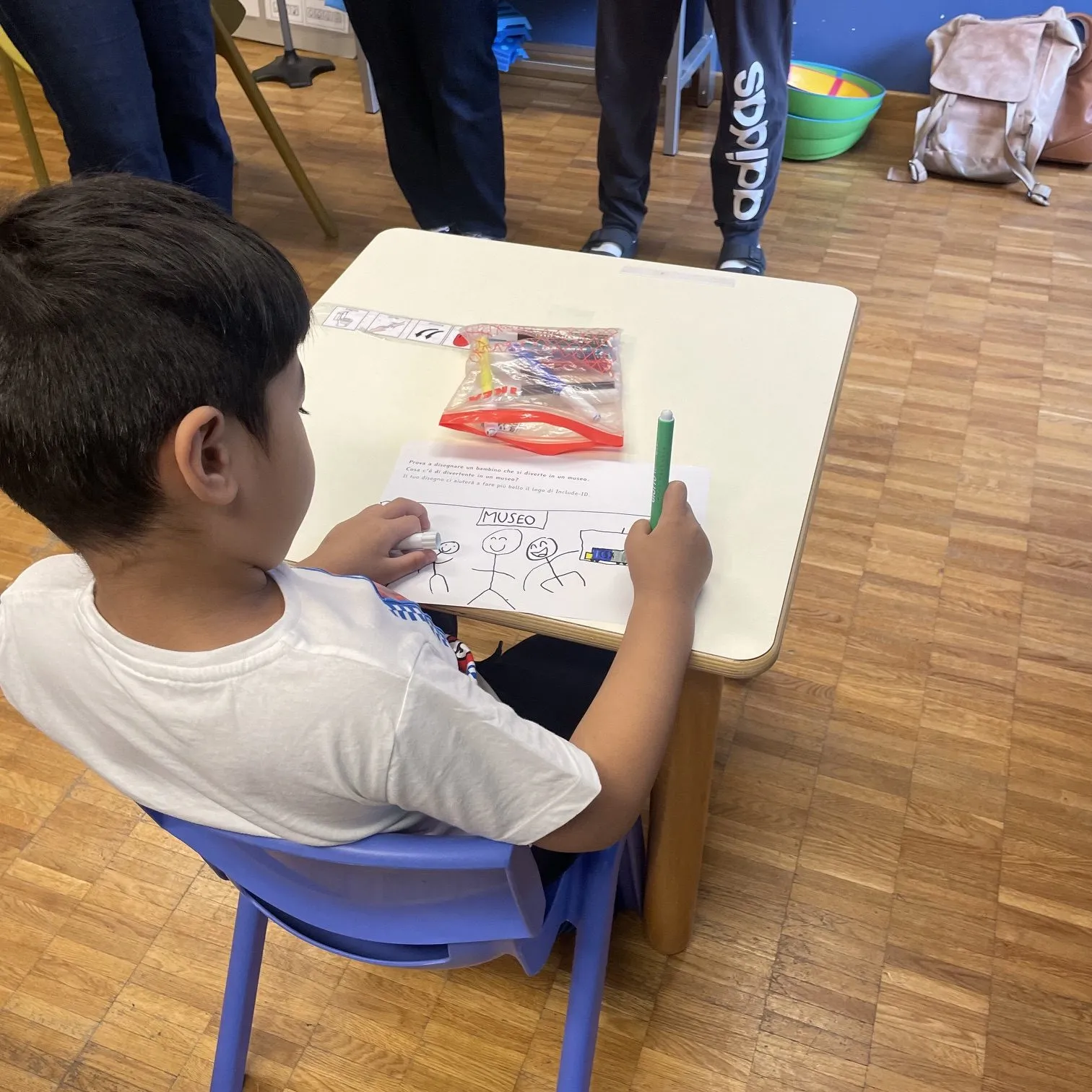 A child draws at a classroom table during the INCLUDE-ID co-design workshop, with picture symbols and coloured markers